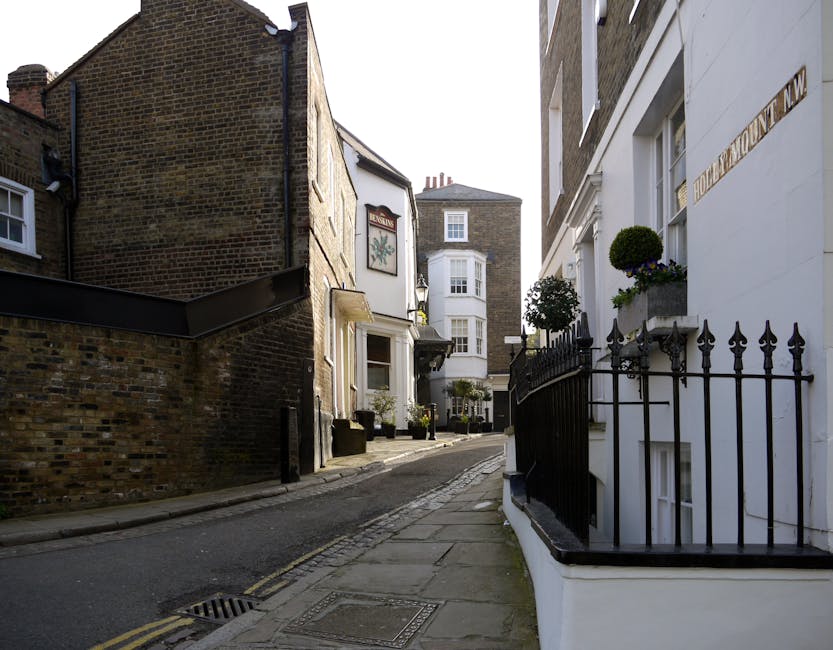 A narrow, paved alleyway in an urban area with a gentle curve, flanked by multi-story buildings on both sides. On the left, a brick wall extends alongside structures with small windows and a sign, whi